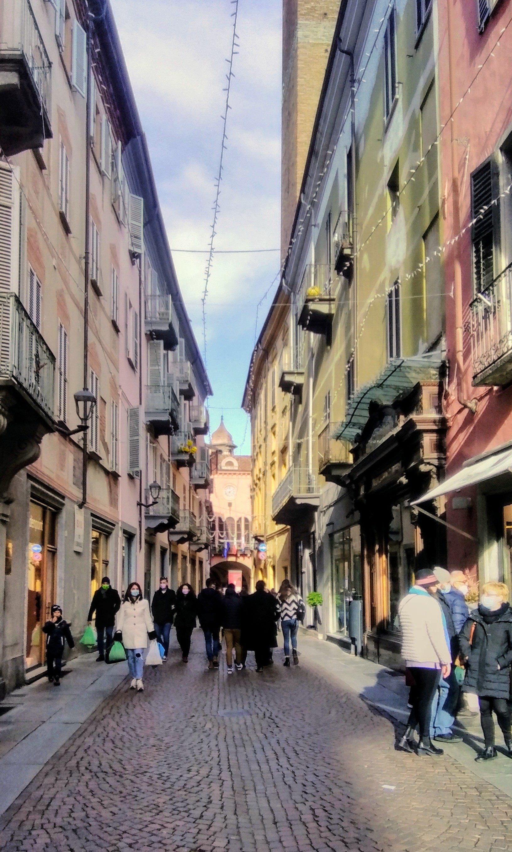 Pessoas caminhando pela Via Vittorio Emanuele, entre fachadas coloridas, com o Palazzo Comunale ao fundo