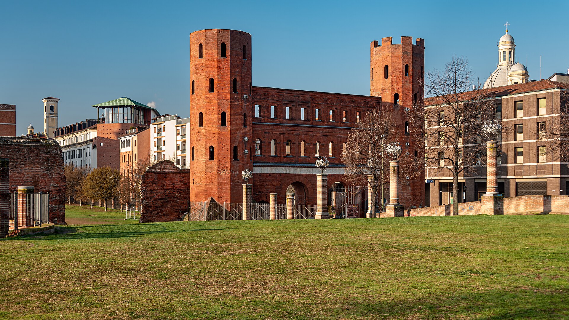 Duas torres de tijolos da Porta Palatina, ligadas por uma fachada com arcos, diante de um gramado em Turim.