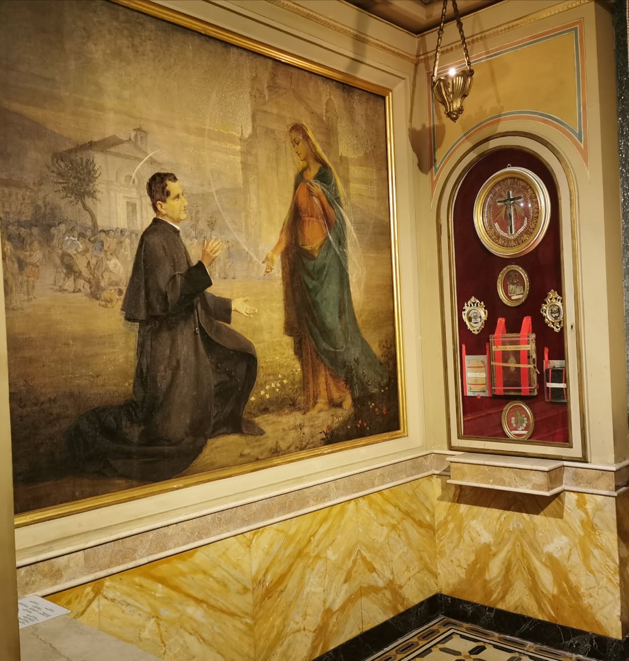 Pintura do encontro de Dom Bosco com Maria junto a uma vitrine de relíquias