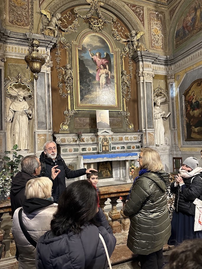 Grupo de visitantes reunido diante do altar da Capela do Anjo da Guarda, ouvindo um homem que aponta para o local
