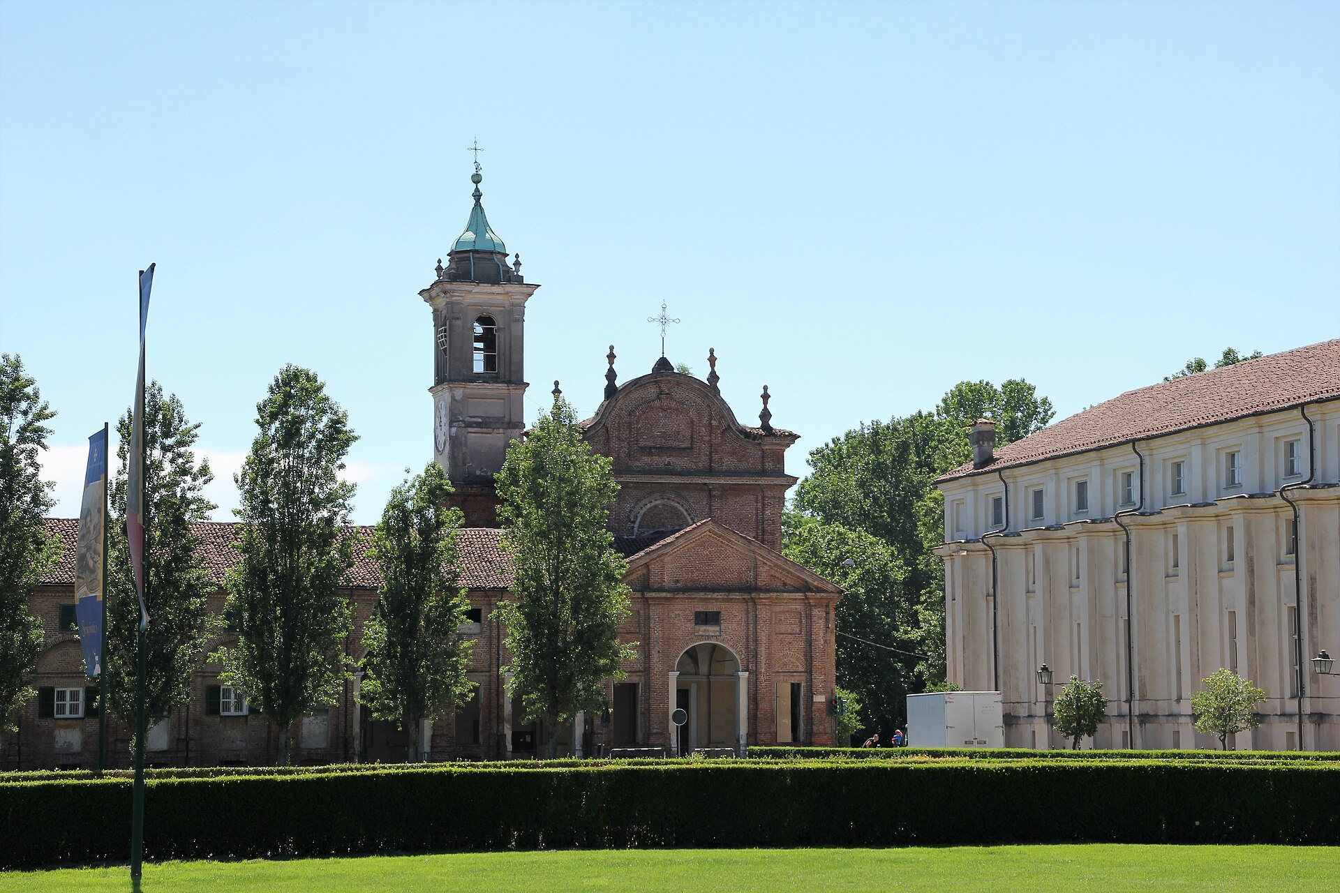 Igreja da Visitazione em Stupinigi, com fachada de tijolos, campanário e árvores, ao lado de um edifício claro.