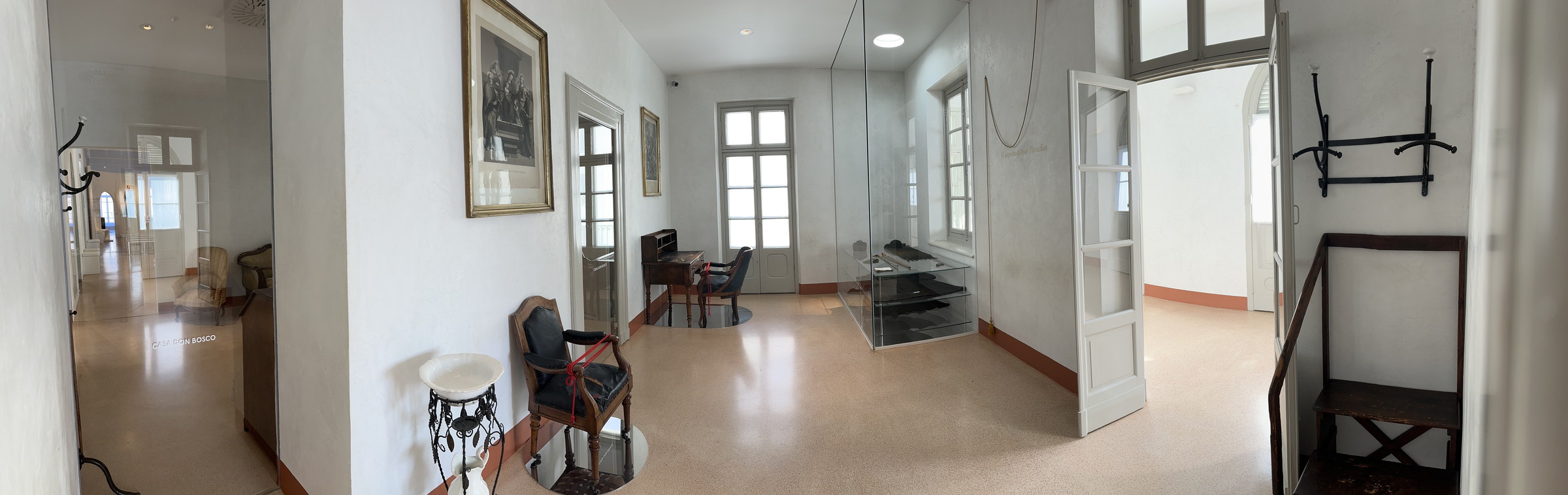 Ambientes claros do Museu Casa Dom Bosco, com portas, janelas, cadeiras, uma escrivaninha e objetos protegidos por vidro.
