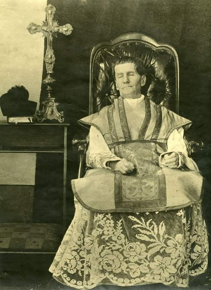 Fotografia histórica de Dom Bosco após a morte, sentado com vestes sacerdotais diante de um crucifixo