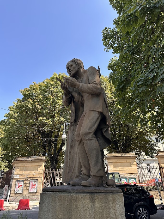 Monumento a Giuseppe Cafasso com a figura de um condenado segurando uma cruz, visto de baixo entre árvores e céu azul, em Turim.