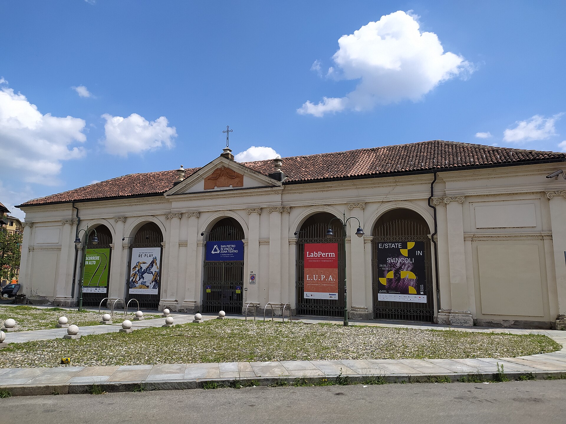 Fachada clara de San Pietro in Vincoli, com aberturas em arco, frontão triangular e cruz, vista da rua sob céu azul.