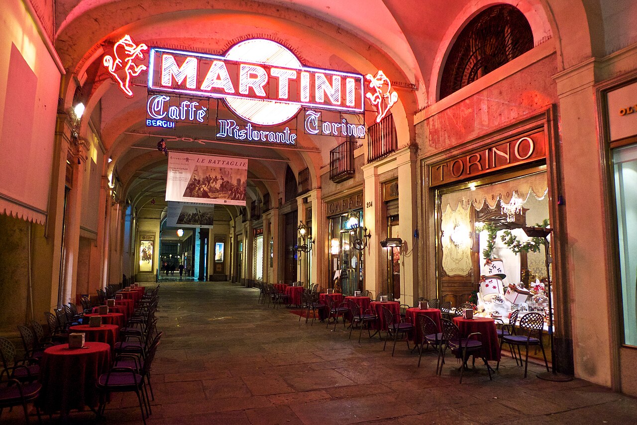 Mesas de café sob os pórticos da Piazza San Carlo, com a fachada do Caffè Torino e uma placa luminosa Martini