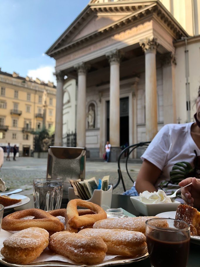 Mesa com café e doces ao ar livre, diante de uma igreja com colunas em Turim